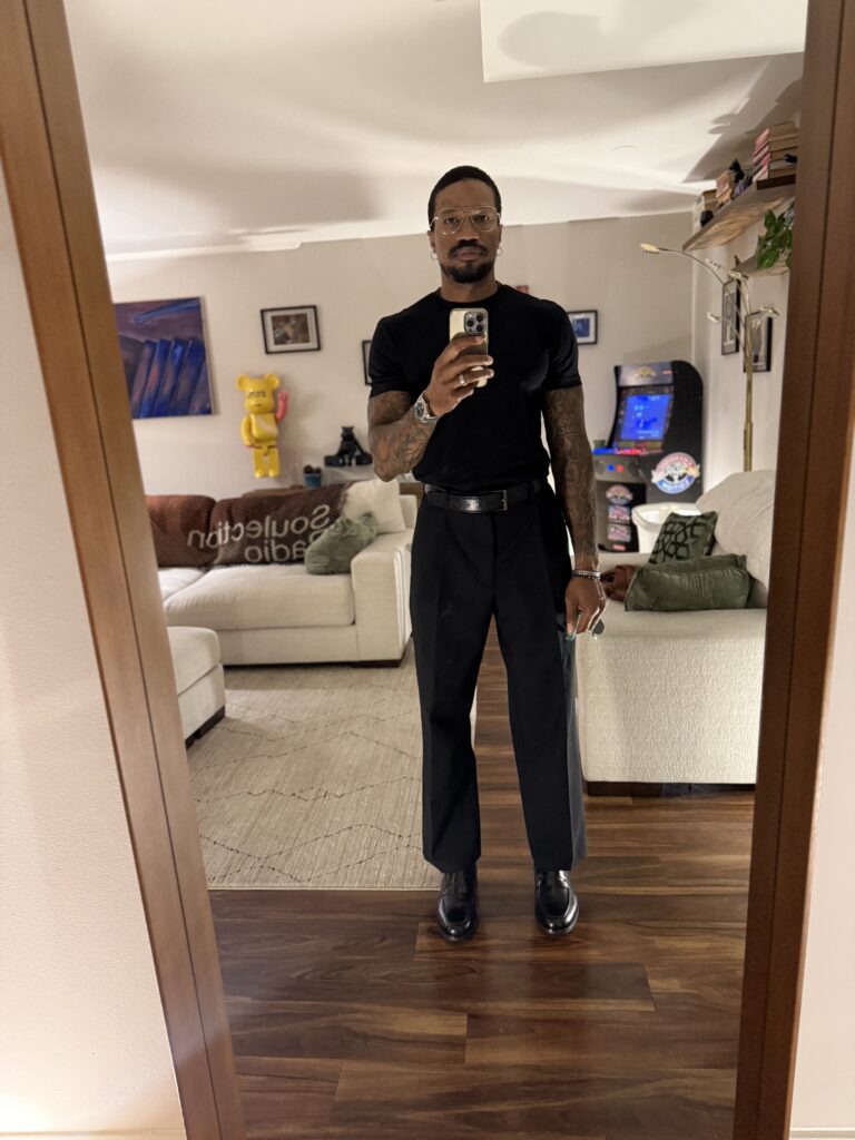Brandon RaSean wearing a fitted black T-shirt, wide-leg black trousers, a belt, and black boots.